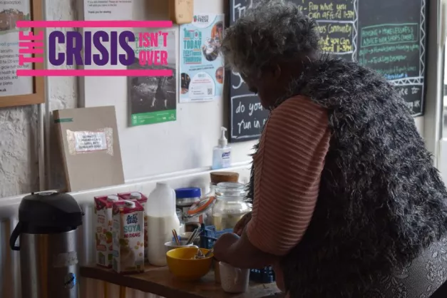 A woman serving breakfast in one of our community living rooms. The image is branded with the text "The crisis isn't over"