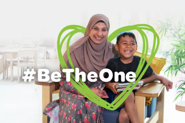 An image of a foster carer and child smiling on a chair, with a green heart graphic and #BeTheOne written over them. 