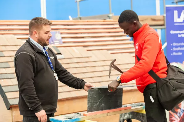 Photo of two men looking at a lead sheeting demonstration. The man on the right is holding on to a tool