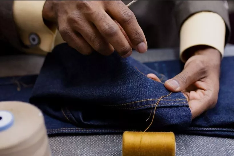 Two hands are shown with needle and thread repairing jeans