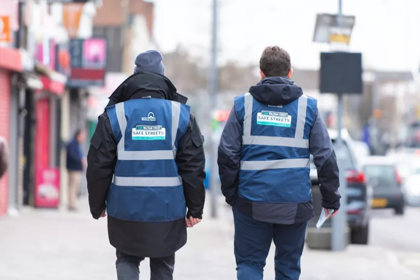 Two male council officers wearing high vis blue jackets that say 'Safe Streets' walking down the street with their backs to camera