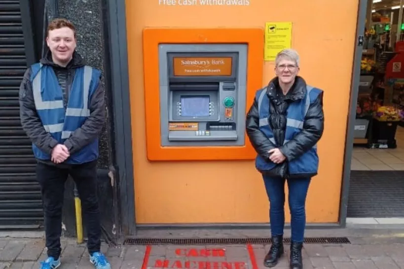 Two Safe Streets officers in high vis blue vests stand outside a Sainsburys ATM, smiling at the camera. On the floor, a privacy box has been spray painted in red paint.
