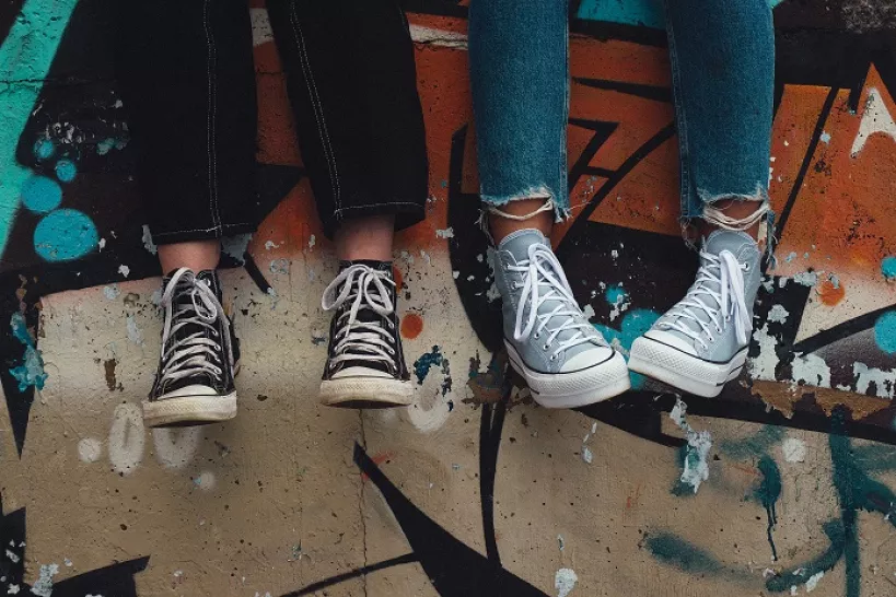 Two teenagers' legs dangle over a wall, both wearing jeans and converse trainers. The wall has bright graffiti on. One person is in black jeans with black trainers, and the other person is in blue jeans with light blue-grey trainers.