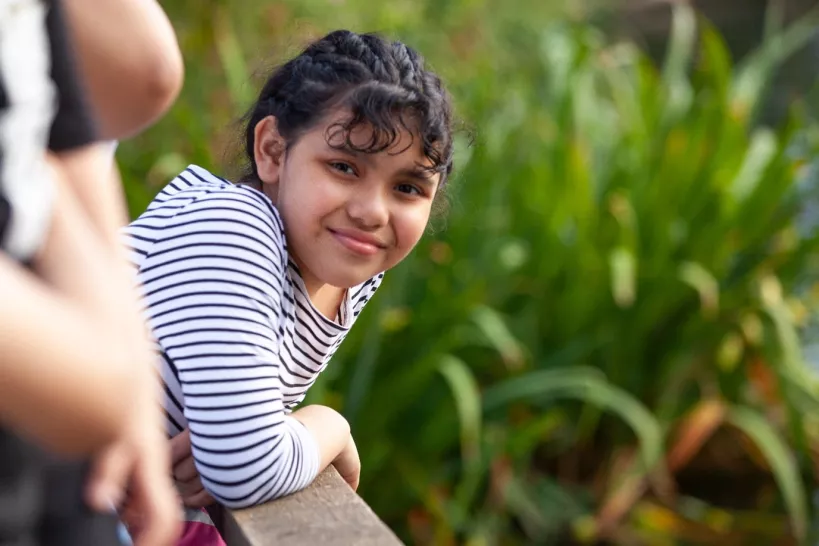 Picture of a young girl smiling and leaning on a wooden fence with tall grass in the background.
